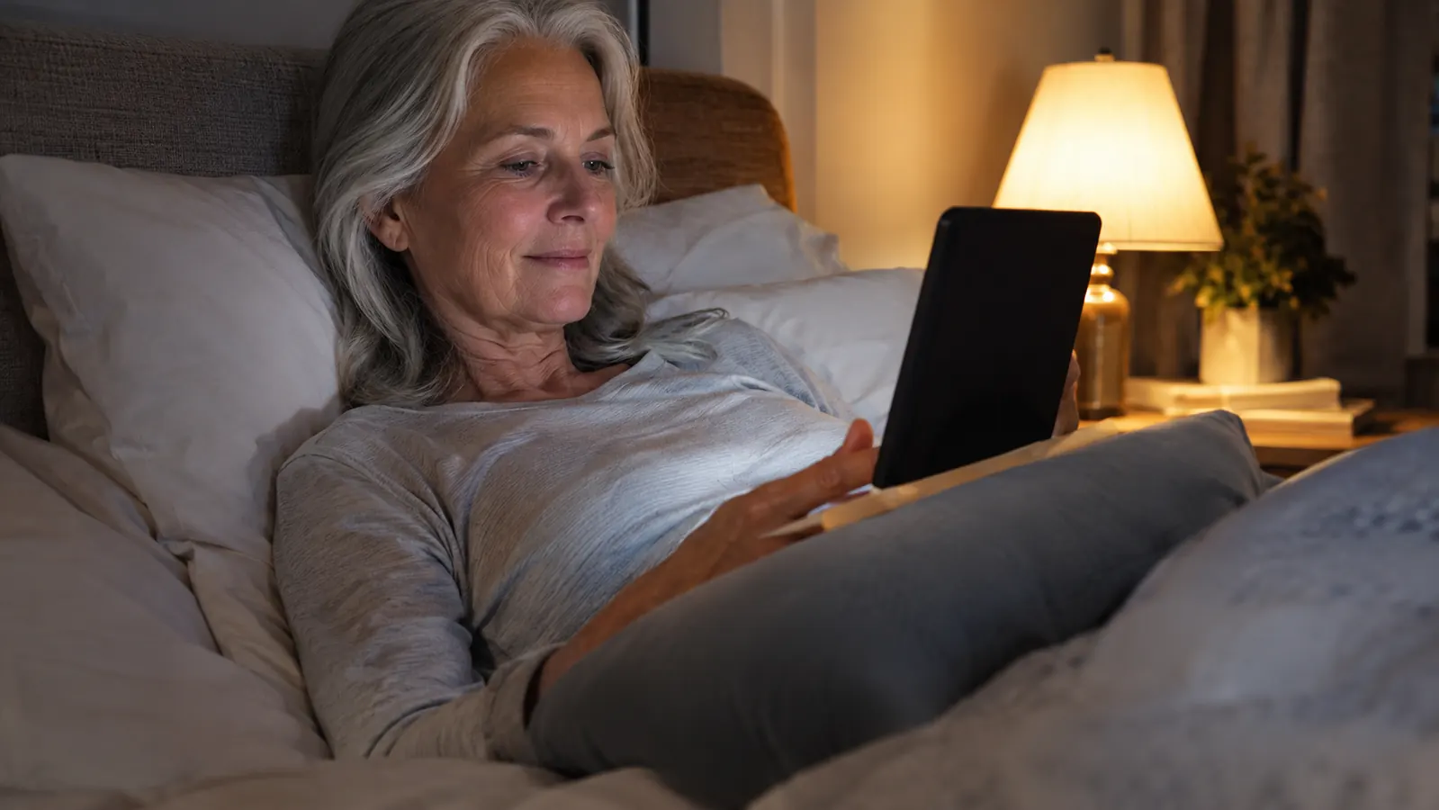 A woman in her sixties reading an e-reader in bed at night with warm lamp light